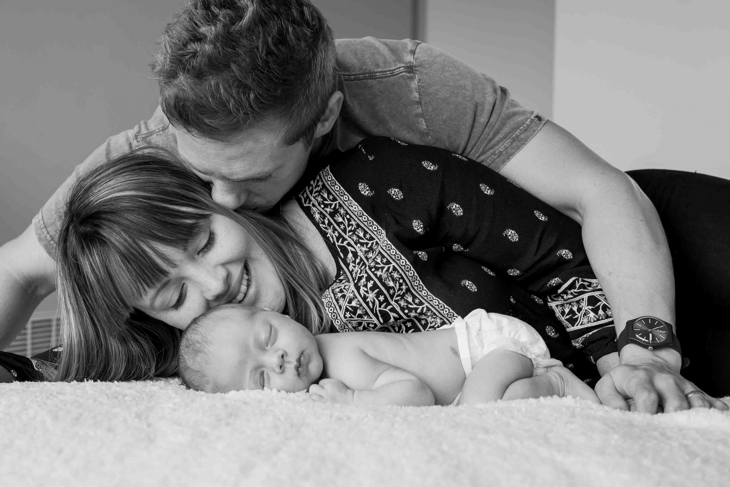 newborn films and photos: newborn sleeping with belly down, mummy laying next to baby looking at him while daddy is kissing her cheek